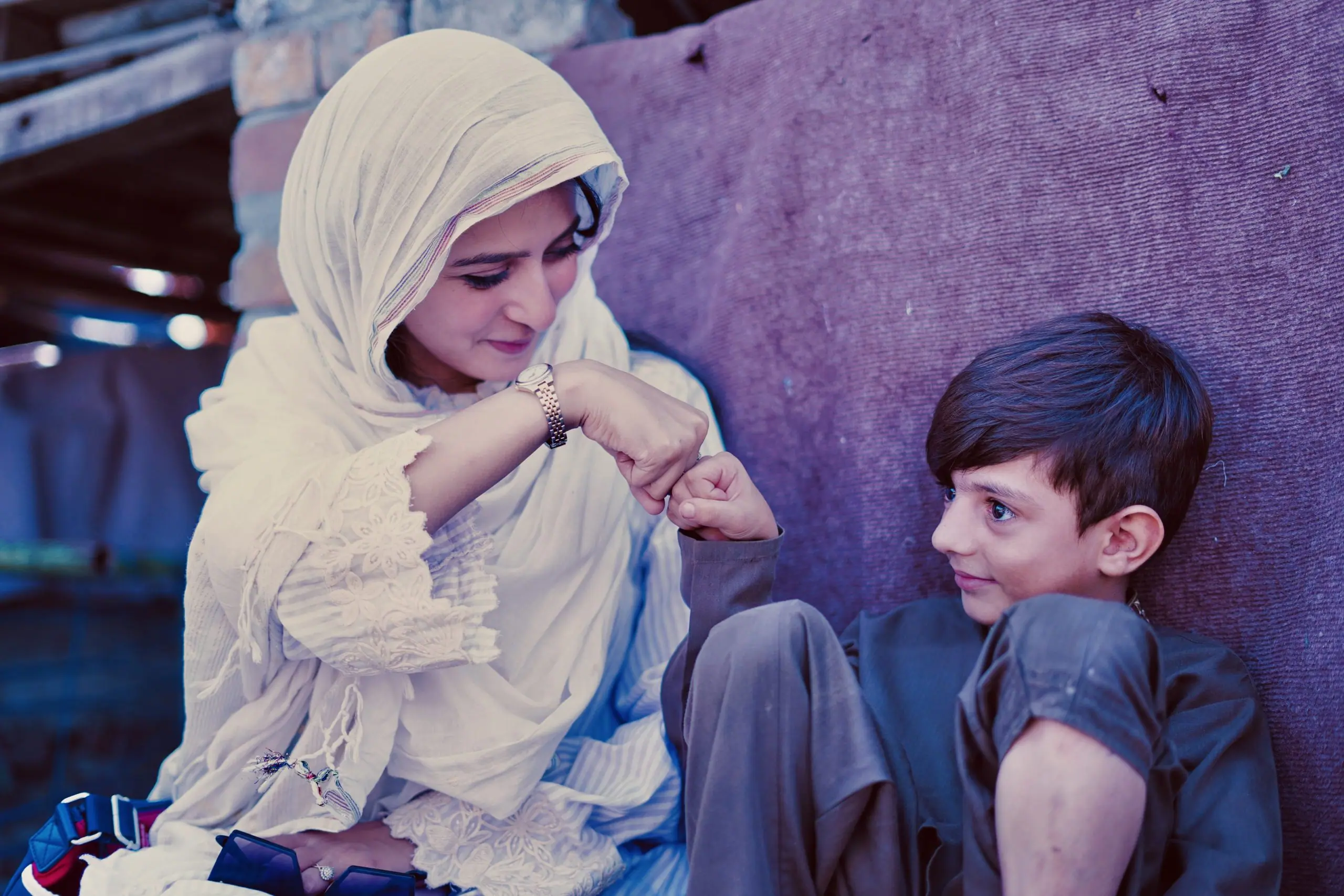 A woman in a cream headscarf and a young boy sit against a wall, smiling as they share a fist bump.